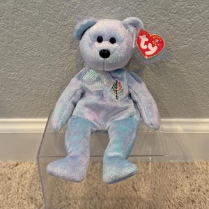 May include: A light blue plush bear with a white ribbon and a small Christmas tree design on its chest. The bear has black eyes and a red heart-shaped tag attached to its ear. The bear is sitting on a clear acrylic display.