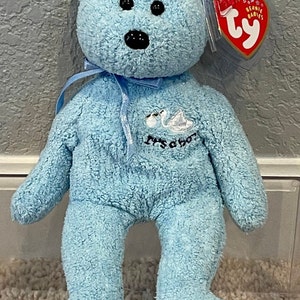 May include: A light blue Beanie Baby plush bear with a white swan embroidered on its chest. The bear has black eyes and a black nose. The tag reads "Beanie Babies" and has a red and white Ty logo.