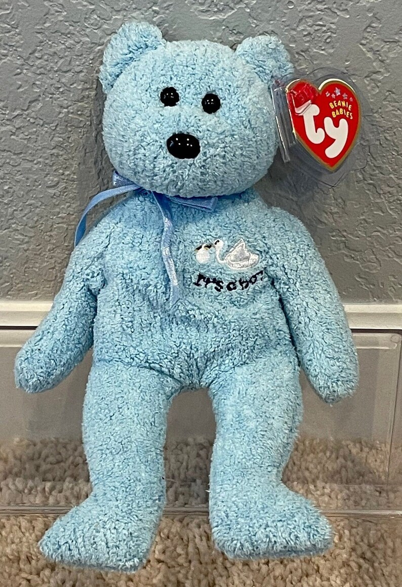 May include: A light blue Beanie Baby plush bear with a white swan embroidered on its chest. The bear has black eyes and a black nose. The tag reads "Beanie Babies" and has a red and white Ty logo.