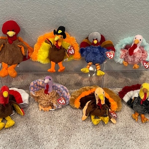 May include: A collection of nine plush turkey toys. The turkeys are all different colors and have different designs. Some of the turkeys have hats on their heads. The turkeys are all sitting on a white surface.