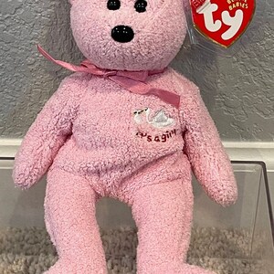 May include: Pink Beanie Baby plush bear with a white swan embroidered on its chest. The bear has a pink ribbon around its neck and a Ty Beanie Babies tag attached to its ear. The tag is red with white stars and the words "Beanie Babies" printed on it.