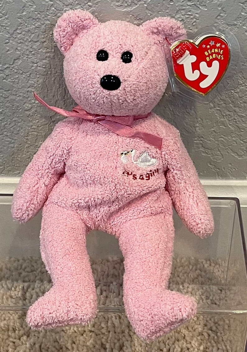 May include: Pink Beanie Baby plush bear with a white swan embroidered on its chest. The bear has a pink ribbon around its neck and a Ty Beanie Babies tag attached to its ear. The tag is red with white stars and the words "Beanie Babies" printed on it.