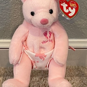May include: Pink Beanie Baby bear wearing a diaper with a white background and pink footprints and the words "New Baby" in pink. The bear has black eyes and a pink nose. The bear has a Ty tag that says "Original Beanie Babies".