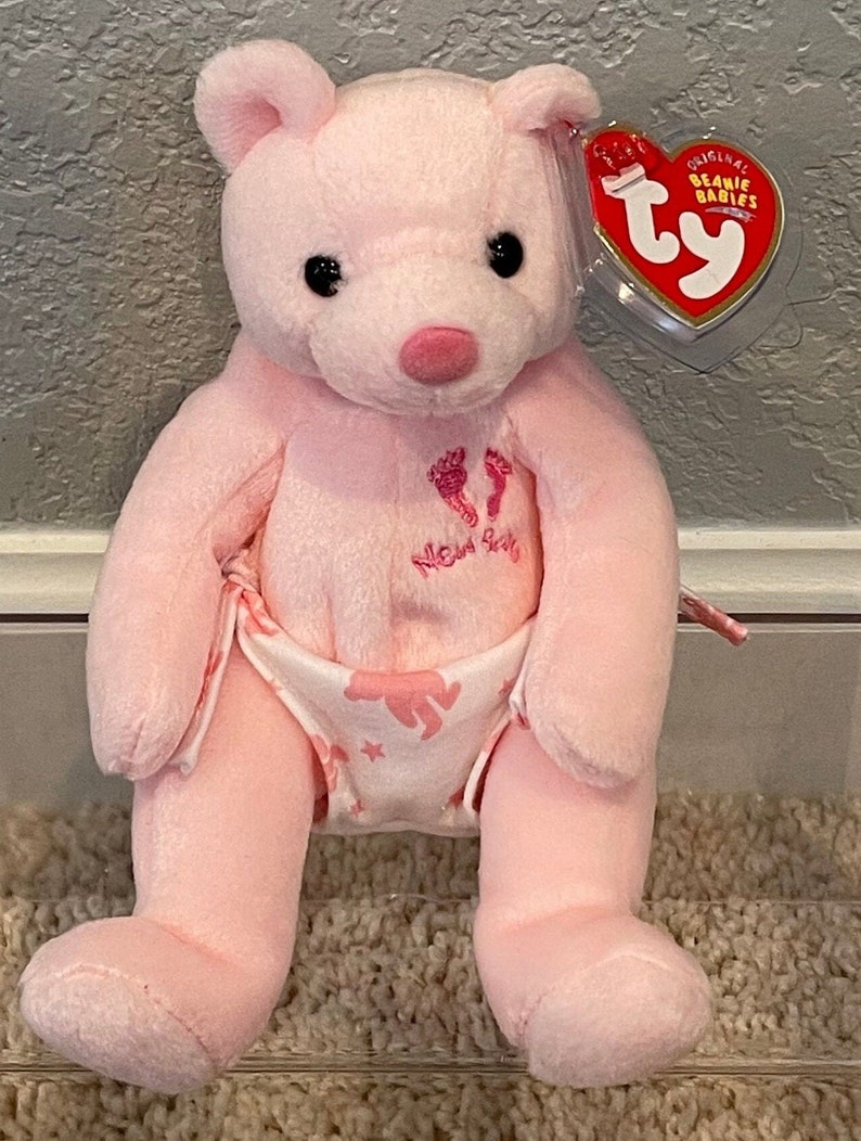 May include: Pink Beanie Baby bear wearing a diaper with a white background and pink footprints and the words "New Baby" in pink. The bear has black eyes and a pink nose. The bear has a Ty tag that says "Original Beanie Babies".