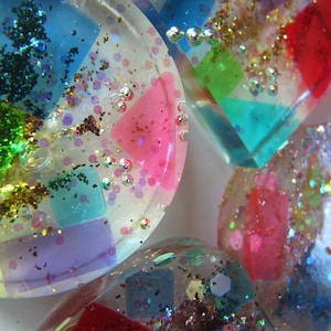 May include: Close-up of several clear resin pieces with colorful geometric shapes and glitter. The pieces feature red, pink, blue, green, and purple squares and triangles, with silver and gold glitter accents.