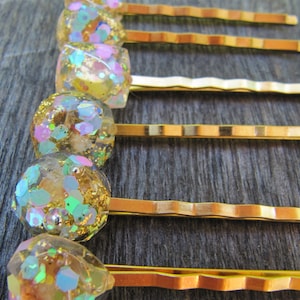 May include: Close-up of several gold-toned bobby pins with decorative tops. The tops are clear resin filled with iridescent glitter in shades of gold, pink, and blue. The bobby pins are arranged vertically, with some straight and some wavy.