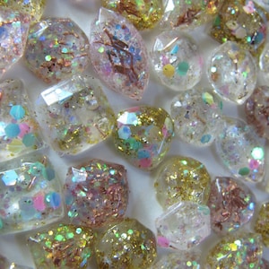May include: A collection of clear, faceted resin gems filled with iridescent glitter in various colors, including gold, pink, and blue. The gems are in various shapes and sizes, creating a sparkling effect.
