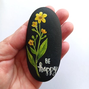 May include: A hand-painted black stone with a yellow flower design and green leaves. The words "BE happy" are written in white script. The stone is oval-shaped and held in a person's hand.