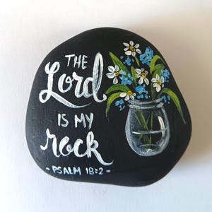 May include: A smooth black river rock painted with white hand-lettered text that reads The Lord is my rock Psalm 18:2. To the right of the text is a painted glass jar containing a bouquet of white daisies and small blue flowers with green leaves.