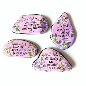 May include: Four smooth, flat stones painted with a light purple watercolor wash and decorated with white flowers and green leaves. Each stone features black hand-lettered Bible verses: Psalm 18:2, Psalm 118:6, Hebrews 13:5, and Matthew 19:26.