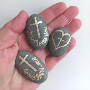 May include: Three gray river stones with gold lettering and designs. Two stones have a cross and the words "Jesus loves you" and "Jesus loves me". The third stone has a heart with a cross inside. Religious themed decor.