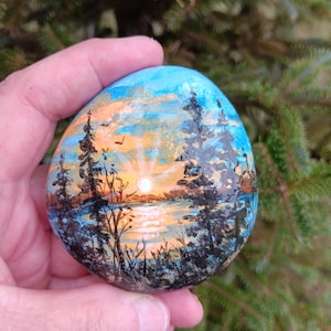 May include: A smooth, rounded river stone hand-painted with a landscape scene featuring a bright sun rising or setting over a calm body of water. The painting includes dark silhouettes of tall pine trees and grasses against a sky of blue and orange.