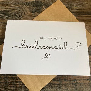 May include: White card with black text that reads "Will you be my bridesmaid?" with a small heart drawn below the text.