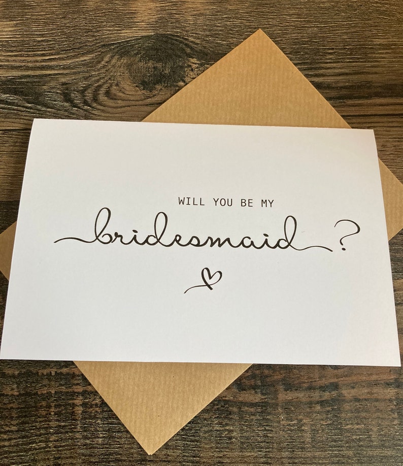May include: White card with black text that reads "Will you be my bridesmaid?" with a small heart drawn below the text.