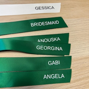 May include: Ribbons with printed text. One white ribbon reads "GESSICA". Several green ribbons display "BRIDESMAID", "ANOUSKA", "GEORGINA", "GABI", and "ANGELA" in white. The ribbons are arranged on a wooden surface.