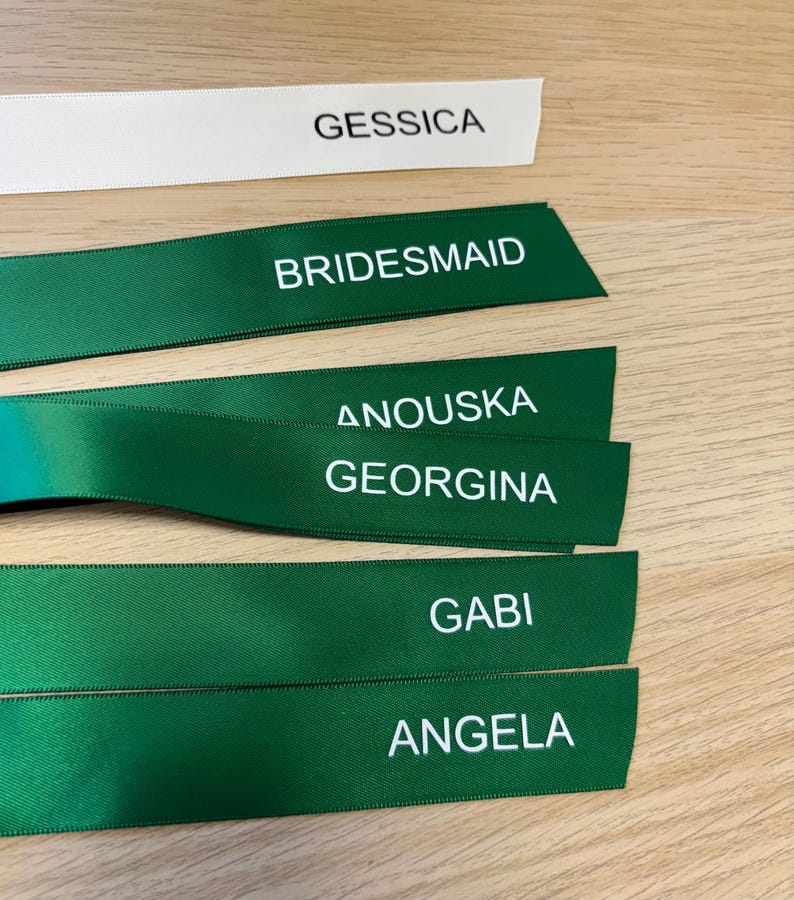 May include: Ribbons with printed text. One white ribbon reads "GESSICA". Several green ribbons display "BRIDESMAID", "ANOUSKA", "GEORGINA", "GABI", and "ANGELA" in white. The ribbons are arranged on a wooden surface.