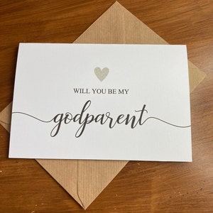May include: A white card with a silver glitter heart and the text "Will you be my godparent" in black cursive writing.