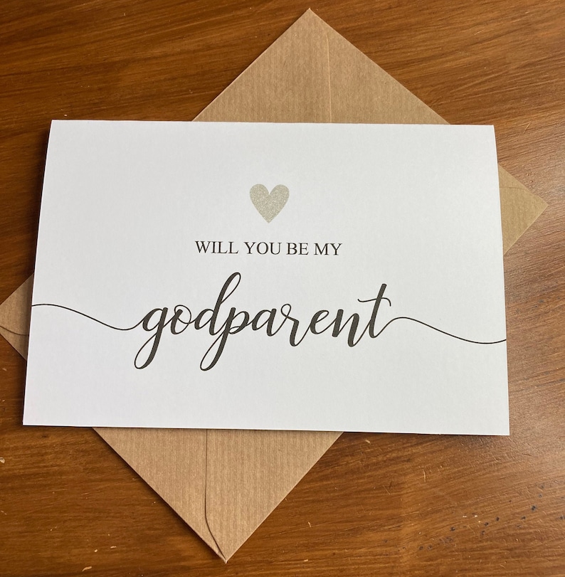 May include: A white card with a silver glitter heart and the text "Will you be my godparent" in black cursive writing.