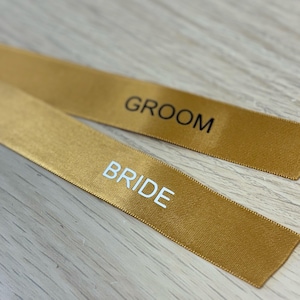 May include: Two gold satin ribbons with the words "BRIDE" and "GROOM" printed in white and black, respectively. The ribbons are lying on a light wood surface. Ideal for wedding decorations.