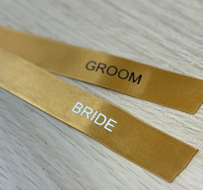 May include: Two gold satin ribbons with the words "BRIDE" and "GROOM" printed in white and black, respectively. The ribbons are lying on a light wood surface. Ideal for wedding decorations.