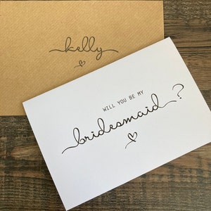 May include: White card with black calligraphy that says "Will you be my bridesmaid?" with a small heart symbol. The card is on top of a brown envelope with the name "Kelly" written on it in black calligraphy.