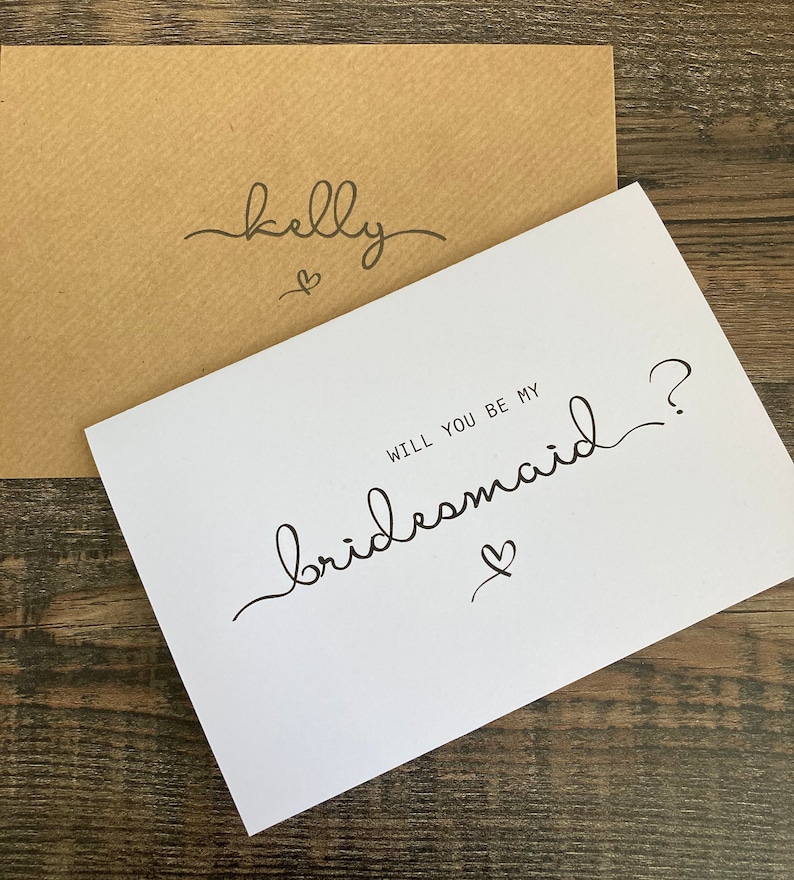 May include: White card with black calligraphy that says "Will you be my bridesmaid?" with a small heart symbol. The card is on top of a brown envelope with the name "Kelly" written on it in black calligraphy.