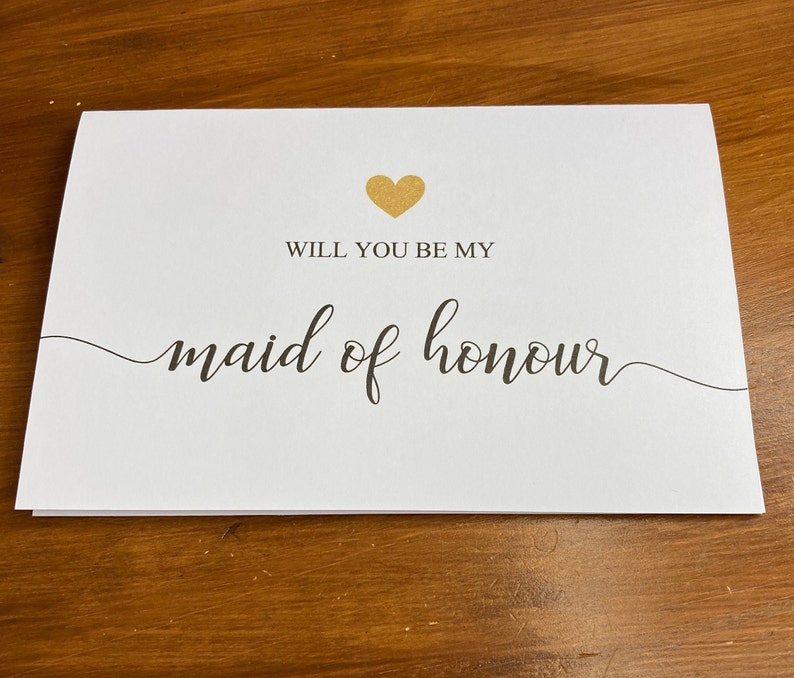 Puede incluir: Tarjeta blanca con un coraz&oacute;n dorado y texto negro que dice "&iquest;Quieres ser mi dama de honor?"