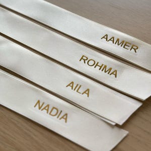 May include: Four ivory satin ribbons with gold lettering. The names "AAMER", "ROHMA", "AILA", and "NADIA" are printed on the ribbons. The ribbons are arranged diagonally, with a light-colored wooden surface in the background.