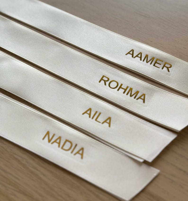 May include: Four ivory satin ribbons with gold lettering. The names "AAMER", "ROHMA", "AILA", and "NADIA" are printed on the ribbons. The ribbons are arranged diagonally, with a light-colored wooden surface in the background.