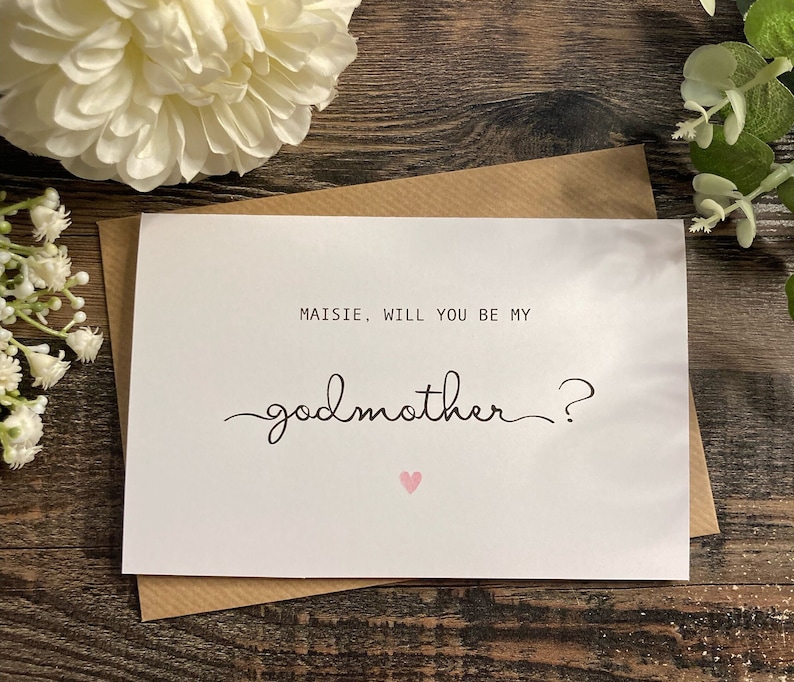 Pu&ograve; includere: Carta bianca con testo nero che dice "Maisie, vuoi essere la mia madrina?" con un cuore rosa sotto il testo.