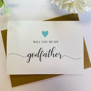 May include: A white card with a blue heart and the text "Will you be my godfather?" in black cursive writing.