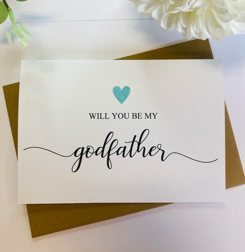 May include: A white card with a blue heart and the text "Will you be my godfather?" in black cursive writing.
