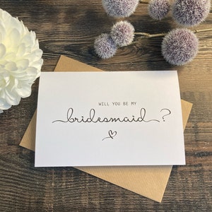 May include: White card with black text that reads "Will you be my bridesmaid?" with a small heart symbol below the text.