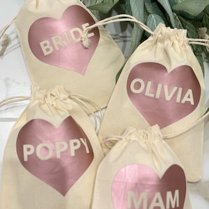 Peut inclure: Quatre sacs en tissu beige avec des cœurs rose doré. Chaque cœur porte un nom ou un mot imprimé : "BRIDE", "OLIVIA", "POPPY" et "MAM". Les sacs sont disposés sur une surface blanche, avec de la verdure en arrière-plan.