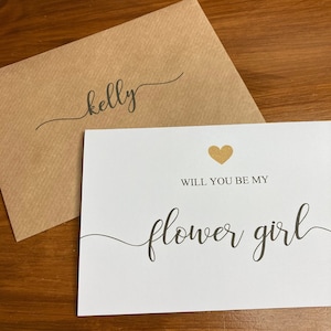 Puede incluir: Una tarjeta blanca con un coraz&oacute;n de purpurina dorada y el texto "&iquest;Quieres ser mi dama de honor?" en escritura cursiva negra. La tarjeta est&aacute; sobre un sobre marr&oacute;n con el nombre "kelly" escrito en escritura cursiva negra.