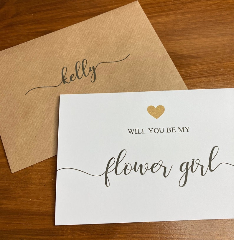 Puede incluir: Una tarjeta blanca con un coraz&oacute;n de purpurina dorada y el texto "&iquest;Quieres ser mi dama de honor?" en escritura cursiva negra. La tarjeta est&aacute; sobre un sobre marr&oacute;n con el nombre "kelly" escrito en escritura cursiva negra.