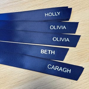 May include: Navy blue satin ribbons with white embroidered names: HOLLY, OLIVIA, BETH, and CARAGH. The ribbons are arranged in a slightly overlapping fan shape. The names are in a simple, sans-serif font.