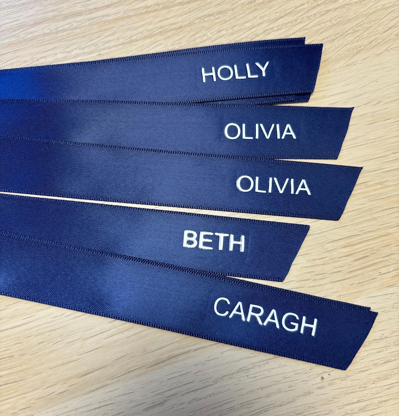 May include: Navy blue satin ribbons with white embroidered names: HOLLY, OLIVIA, BETH, and CARAGH. The ribbons are arranged in a slightly overlapping fan shape. The names are in a simple, sans-serif font.