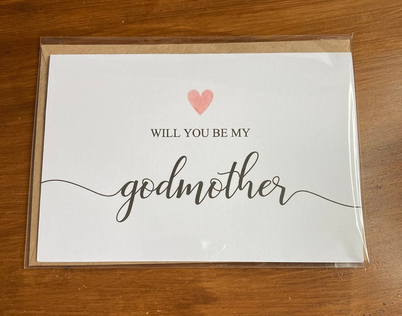 May include: White card with a pink heart and the text "Will you be my godmother" in black cursive writing.