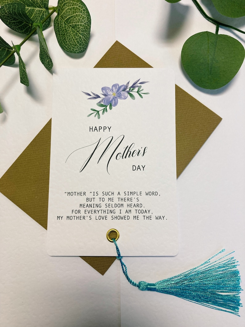 May include: A white card with a purple floral design and the text "Happy Mother's Day" in black script. The card has a gold tassel attached to the bottom and the text "Mother" is such a simple word, but to me there's meaning seldom heard. For everything I am today, my mother's love showed me the way."
