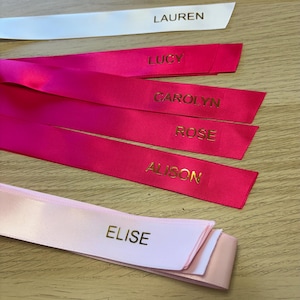 May include: A collection of personalized satin ribbons in various colors. The ribbons are white, pink, and fuchsia, each with a name printed in gold lettering: LAUREN, LUCY, CAROLYN, ROSE, ALISON, and ELISE. The ribbons are arranged on a light wood surface.