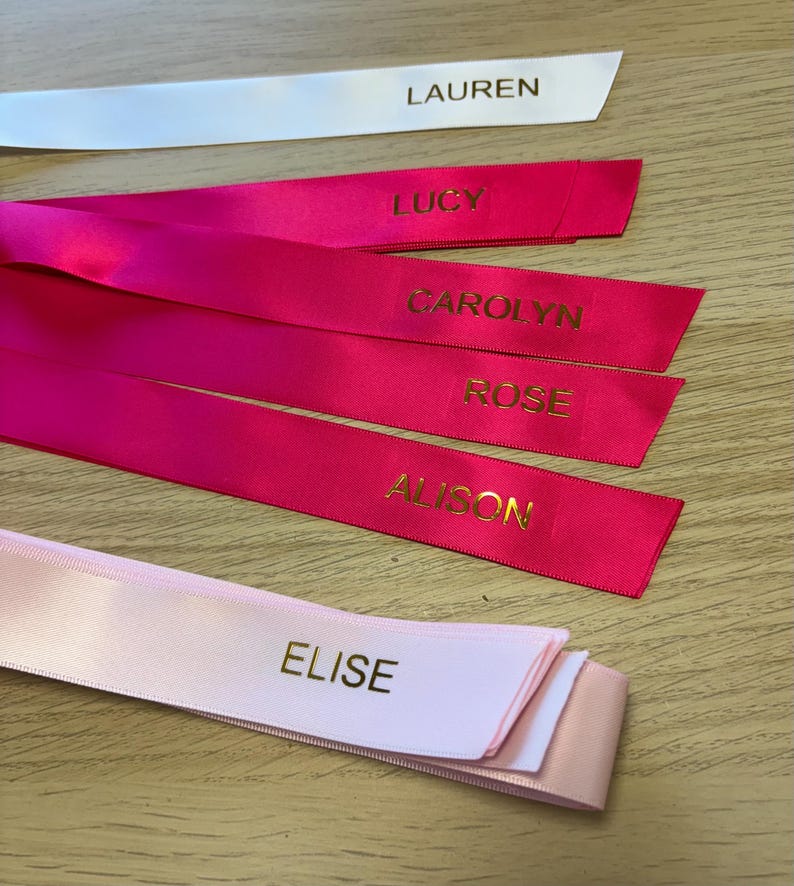 May include: A collection of personalized satin ribbons in various colors. The ribbons are white, pink, and fuchsia, each with a name printed in gold lettering: LAUREN, LUCY, CAROLYN, ROSE, ALISON, and ELISE. The ribbons are arranged on a light wood surface.