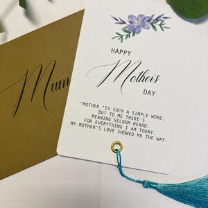 May include: A white card with a blue floral design and the text "Happy Mother's Day". The card has a quote about motherhood and a blue tassel.