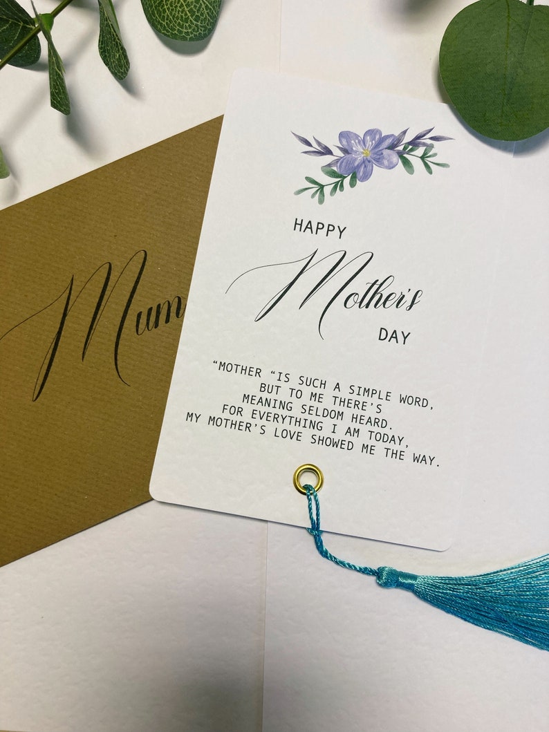 May include: A white card with a blue floral design and the text "Happy Mother's Day". The card has a quote about motherhood and a blue tassel.
