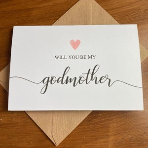 May include: A white card with a pink heart and the text "Will you be my godmother" in black cursive writing.