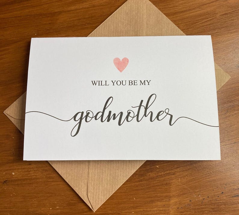 May include: A white card with a pink heart and the text "Will you be my godmother" in black cursive writing.