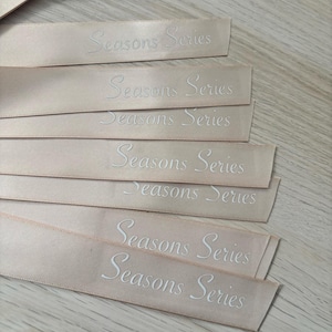 May include: Several pale peach satin ribbons with the words "Seasons Series" embroidered in white. The ribbons are arranged in a slightly overlapping pattern, with the text clearly visible. The ribbons are likely for crafting or gift wrapping.
