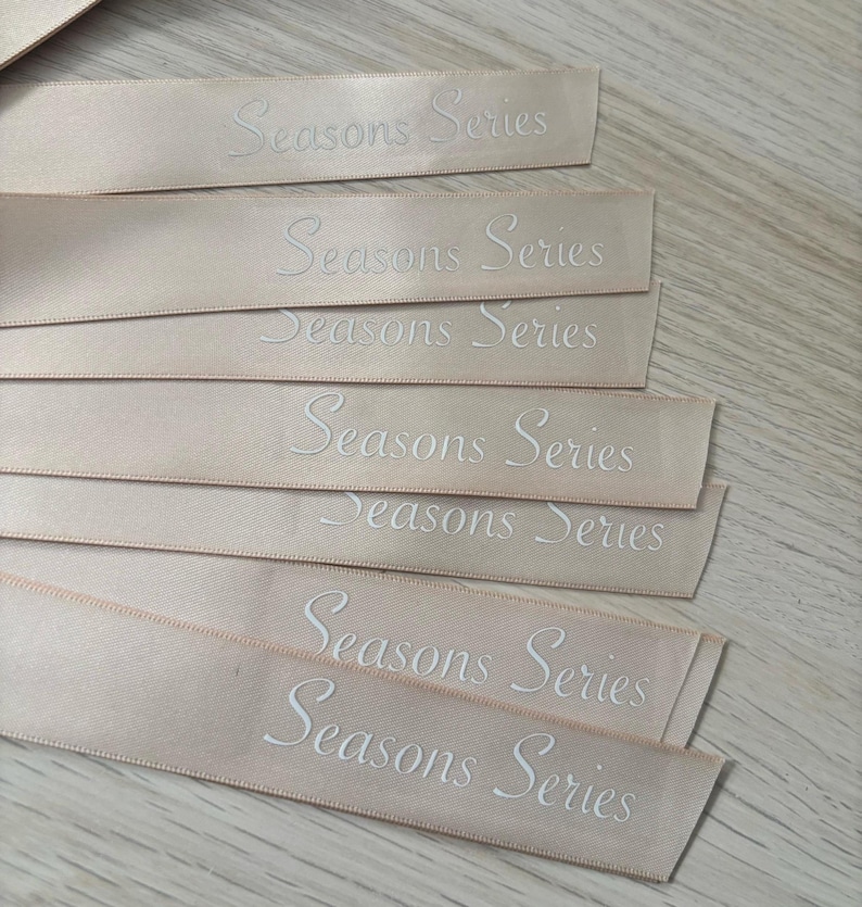 May include: Several pale peach satin ribbons with the words "Seasons Series" embroidered in white. The ribbons are arranged in a slightly overlapping pattern, with the text clearly visible. The ribbons are likely for crafting or gift wrapping.