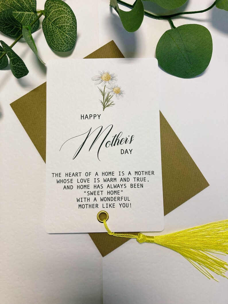 May include: A white card with a yellow tassel attached. The card has a floral design with white daisies and green leaves. The text on the card reads "Happy Mother's Day" and "The heart of a home is a mother whose love is warm and true, and home has always been "Sweet Home" with a wonderful mother like you!"