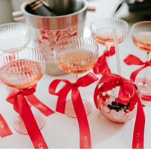 May include: A festive arrangement featuring champagne glasses filled with a pink beverage, adorned with red ribbons. A disco ball-shaped glass and a champagne bucket are also present, creating a celebratory atmosphere. The glasses have a ribbed design.